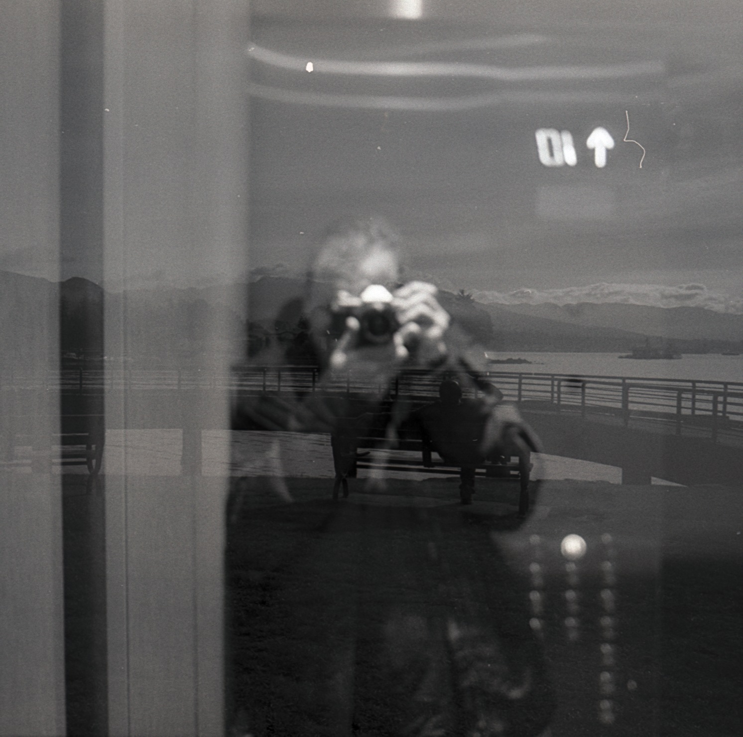 A reflection of the photographer holding a camera, with a waterfront scene behind the glass